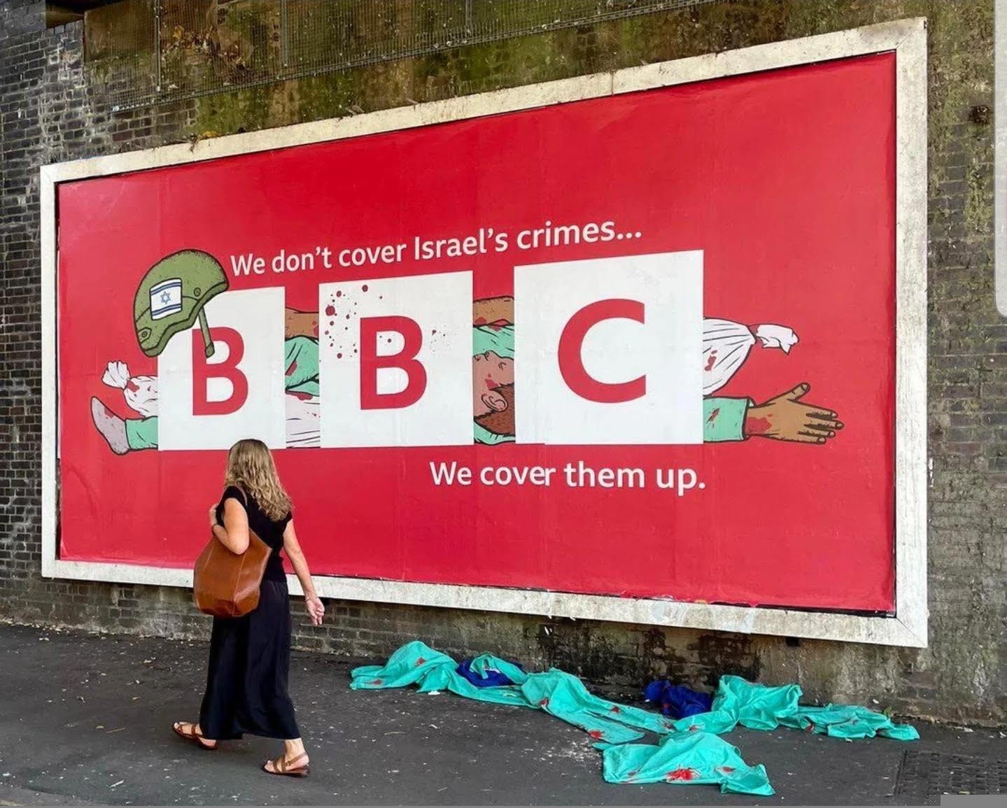 L'immagine mostra un grande cartellone pubblicitario su un muro di mattoni sotto un ponte, di Londra. Il logo della BBC copre il disegno di alcune persone morte, il testo riporta: "We don't cover Israel's crimes... We cover them up."