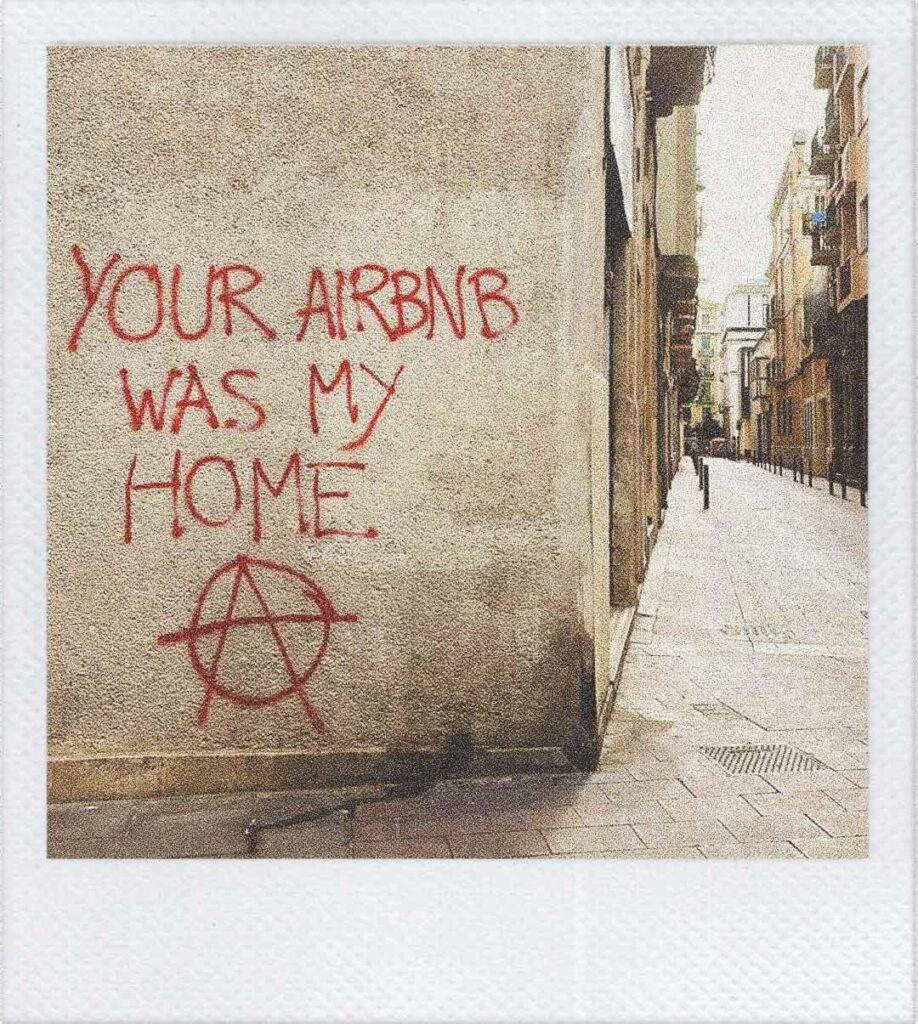 Un muro di Barcellona con una scritta spray rossa: "YOUR AIRBNB WAS MY HOME" e la (A) simbolo dell'anarchia.